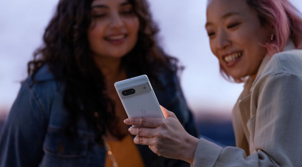 Zwei Frauen gucken draußen bei Dämmerung lächelnd auf ein Smartphone-Dislplay.