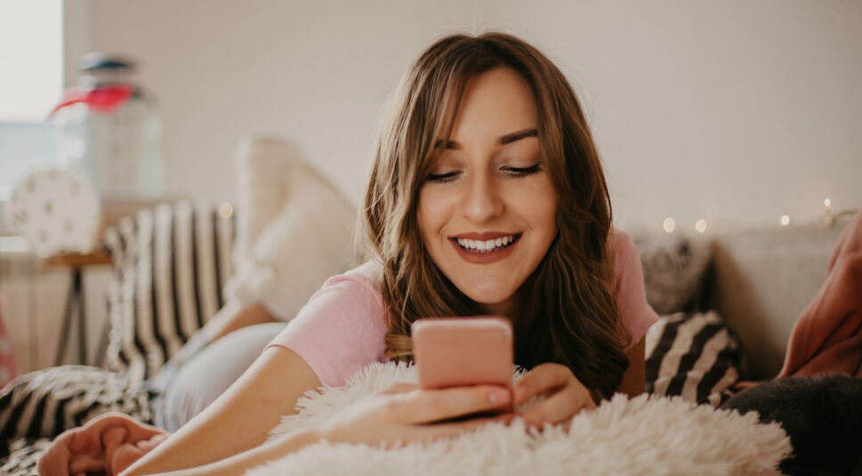 junge Frau liegt auf dem Bett und schaut auf ihr rosa Smartphone