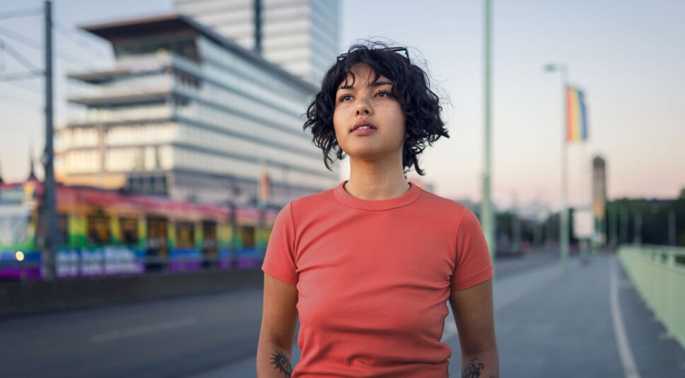 Auf dem Bild zum CSD mit Vodafone steht eine junge Frau auf einer Brücke in Köln. Hinter ihr Fährt eine Bahn in Regenbogenfarben, was das Thema Pride weiter verstärkt.