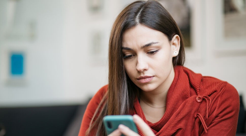 Eine junge Frau mit langen dunklen Haaren und rotem Strickpullover blickt ernst auf ihr türkisfarbenes Smartphone. Der unscharfe Innenraum im Hintergrund lenkt den Fokus auf ihre nachdenkliche, leicht besorgte Mimik. Das Motiv zeigt, wie Nutzer Betrug erkennen und bei einem verdächtigen Ping-Anruf aufmerksam reagieren können, bevor sie zurückrufen oder persönliche Daten preisgeben.