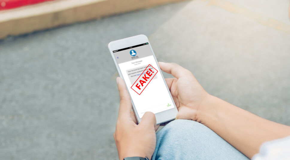 Eine Person hält ein weißes Smartphone in beiden Händen, auf dessen Display eine unscharfe Nachricht mit einem auffälligen roten „FAKE!“-Stempel zu sehen ist. Die Aufnahme verdeutlicht, wie Nutzer Betrug erkennen und verdächtige Inhalte prüfen können. Besonders beim Phishing warnen solche Nachrichten vor gefälschten Absendern, schädlichen Links und dem möglichen Diebstahl persönlicher Daten.
