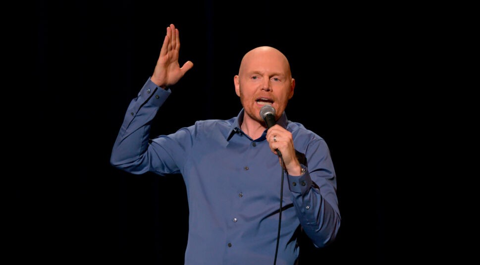 Bill Burr steht auf einer Comedy-Bühne und erhebt seine Hand