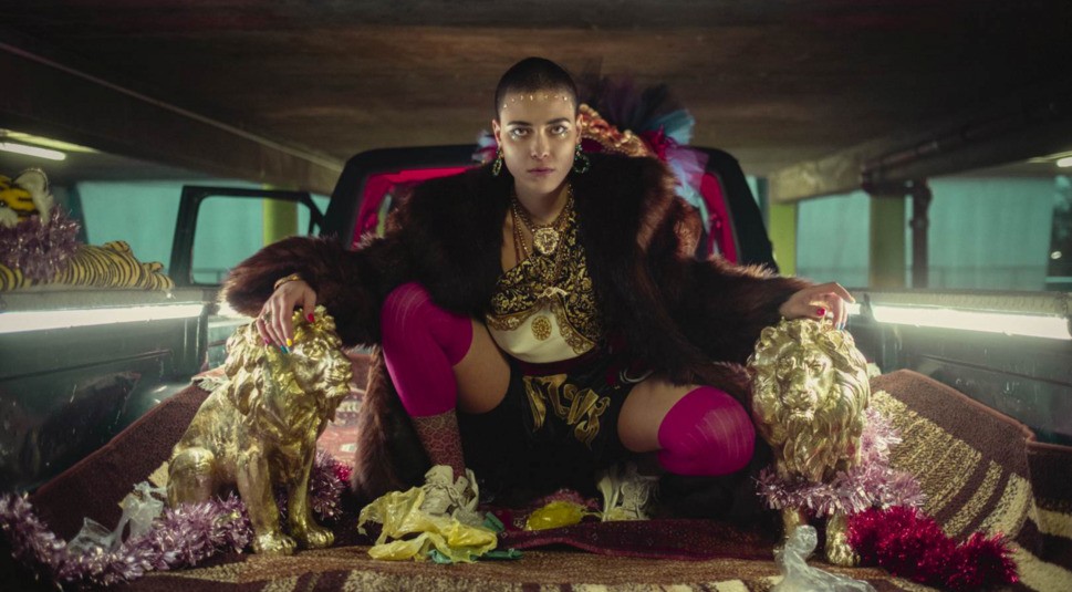 Ein junges Mädchen mit abrasierten Haaren sitzt in einem buntgeschmückten Lieferwagen und posiert für ein Musikvideo zwischen zwei goldenen Löwen-Statuen.