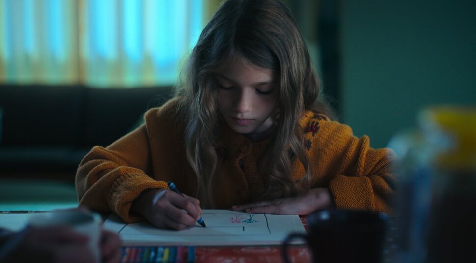 Ein kleines Mädchen mit langen Haaren und einem Strickpulli sitzt an einem Tisch und malt ein Bild