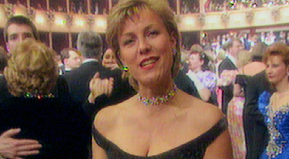 Im Hintergrund tanzen edel gekleidete Personen auf einem Ball, während eine Frau mit kurzen blonden Haaren frontal in die Kamera blickt.