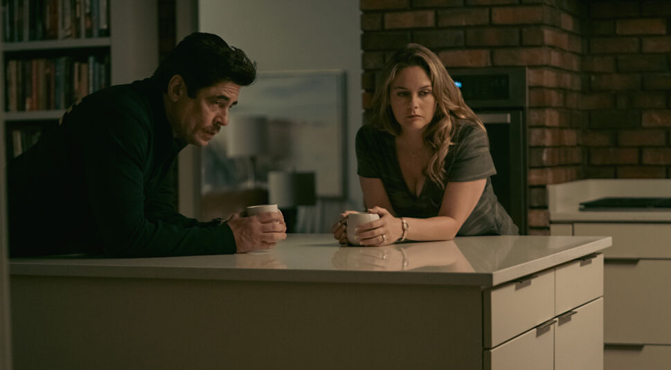 Ein Mann mit dunklen Haaren und Schnauzer und eine blonde Frau lehnen gemeinsam auf einer Kücheninsel.