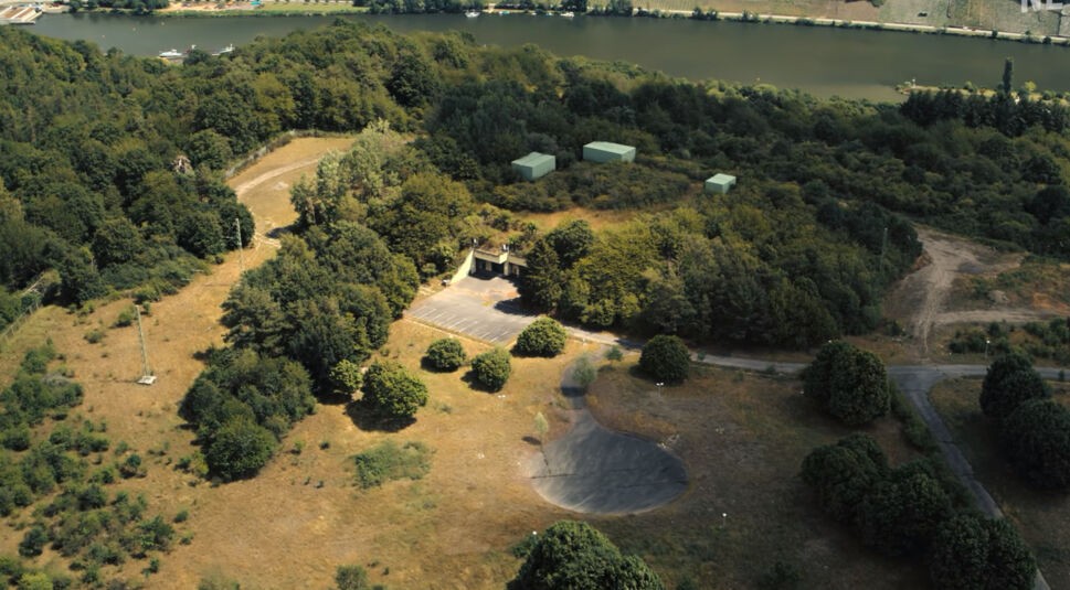 Cyberbunker bei Netflix: Eine umzäunte Bunker-Anlage mit mehreren Gebäuden in einem Waldstück an einem Fluss. 