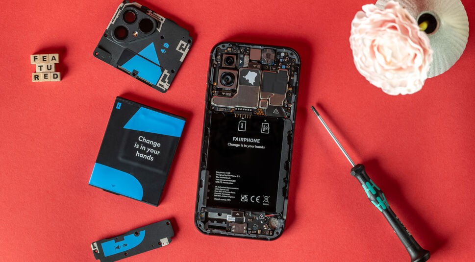 Das Fairphone 5 mit geöffneter Rückseite, Kamera, Speicher und Akku neben einer Blume und Buchstabenwürfeln vor rotem Hintergrund
