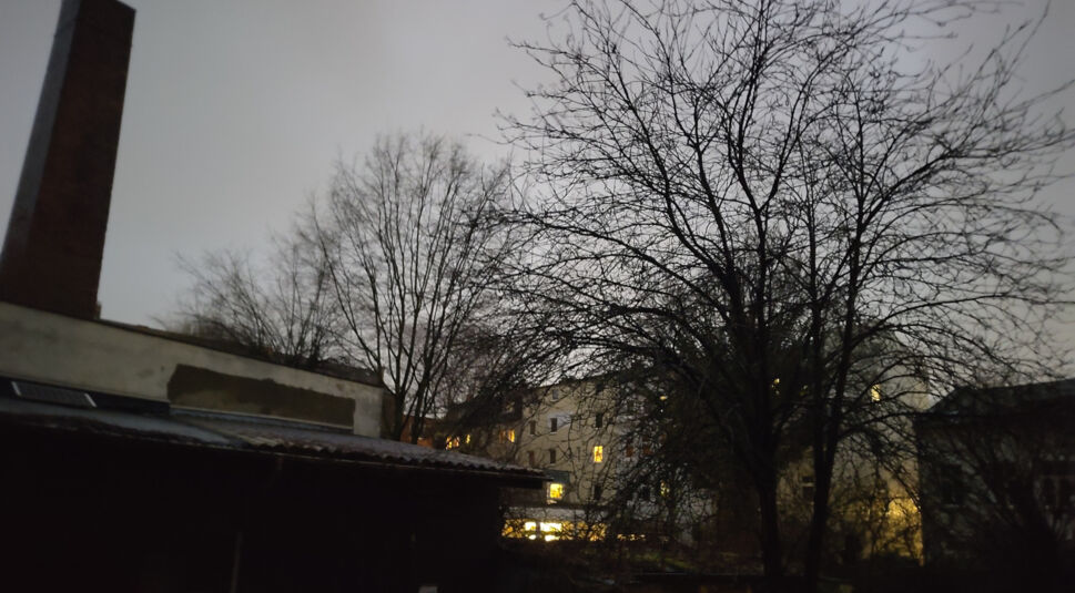 Hinterhof am frühen Morgen mit hellen und dunkeln Bereichen, Fabrikschornstein, Bäumen und einigen erleuchteten Fenstern