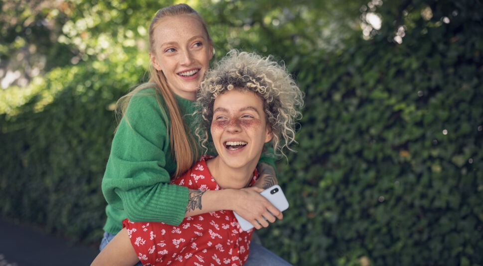 eine Junge Frau hat eine zweite junge Frau huckepack auf dem Rücken, die ein Smartphone in der Hand hält