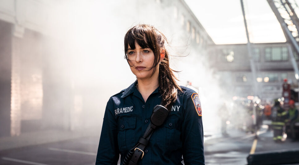 Dakota Johnson in Madame Web:  Eine Frau mit braunen langen Haaren und einer Rettungsdienst-Uniform steht auf einem großen Parkplatz. Im Hintergrund ist ein großes Gebäude und ein Kran zu sehen. 