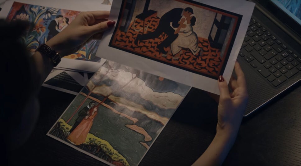 Eine Frau sitzt vor ihrem Laptop und hält mehrere Gemälde in ausgedruckter Form in ihren Händen.