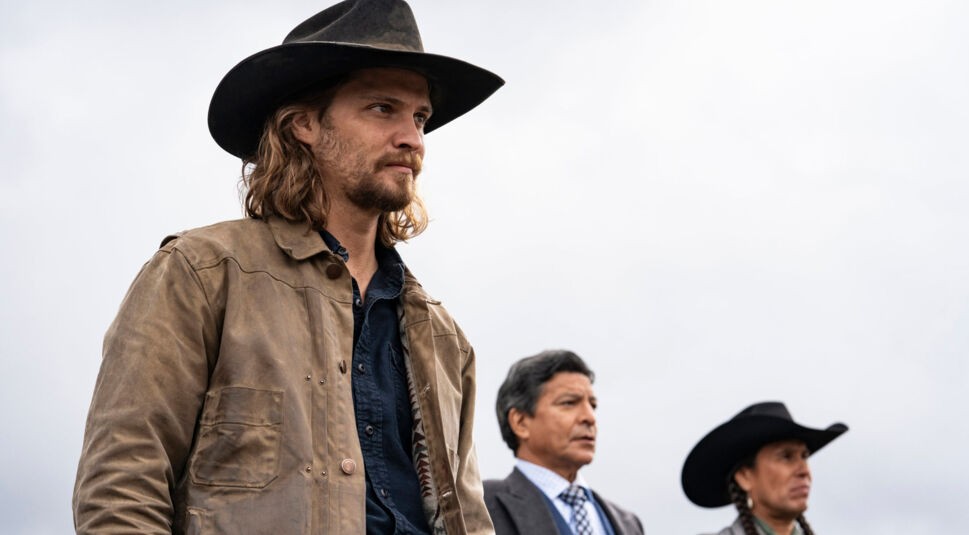 Auf dem Bild zur Fakten Yellowstone Serie sind von links nach rechts Luke Grimes, Gil Birmingham und Moses Brings Plenty zu sehen. Luke Grimes trägt eine braune Lederjacke und einen schwarzen Cowboyhut, während Gil Birmingham im grauen Anzug mit Krawatte steht. Moses Brings Plenty trägt ebenfalls einen Cowboyhut und ein traditionell inspiriertes graues Outfit. Der Hintergrund ist wolkig, was die Szene ernst und nachdenklich wirken lässt. Keyword: Fakten Yellowstone Serie.