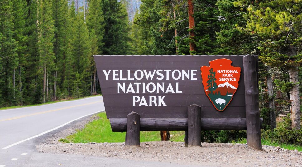 Dieses Bild zum Faktenartikel zur Yellowstone-Seriezeigt das ikonische Eingangsschild des Yellowstone National Parks, umgeben von üppigem Grün und hohen Nadelbäumen. Das braune Holzschild trägt die Aufschrift „Yellowstone National Park“ in weißen Buchstaben und das Emblem des National Park Service mit Berg- und Bisonmotiven. Links führt eine asphaltierte Straße in den Park hinein.