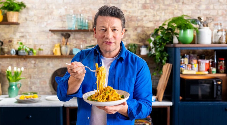 Koch Jamie Oliver trägt ein blaues Hemd und zeigt sein Pasta-Gericht in die Kamera