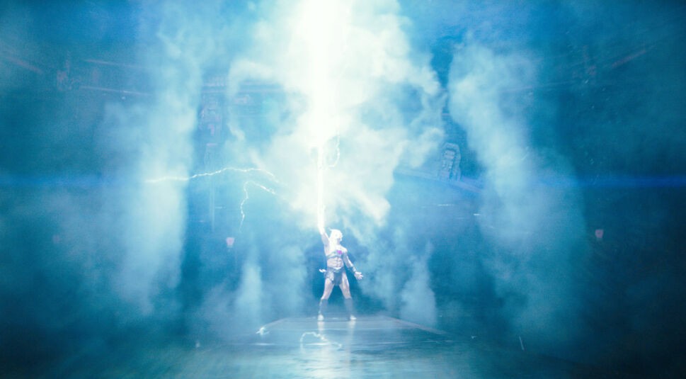 Im Bild zum Masters of the Universe neuer Film steht He-Man, gespielt von Nicholas Galitzine, auf einer Plattform und streckt ein leuchtendes Schwert in den Himmel. Ein greller Blitz durchzuckt die Szene, umgeben von dichten Rauchwolken und magischer Energie. Die Pose ist ikonisch und symbolisiert Macht und Transformation.