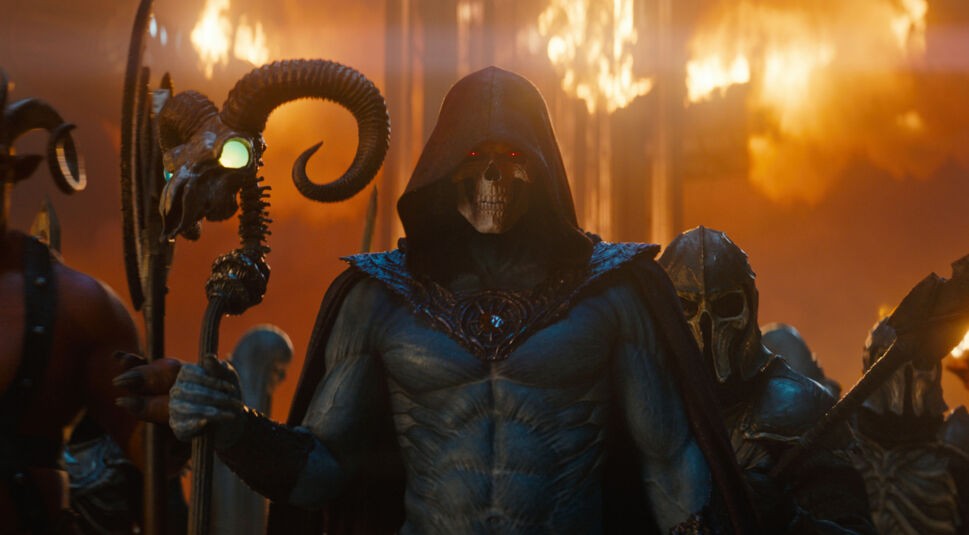 Im Bild zum Masters of the Universe neuer Film ist Skeletor, gespielt von Jared Leto, in bedrohlicher Pose zu sehen. Mit leuchtend roten Augen, Kapuze und Totenschädel-Gesicht hält er einen Stab mit leuchtendem Kristall. Hinter ihm lodern Flammen, umgeben von dunklen Kriegern – die Szene wirkt düster, mächtig und apokalyptisch.