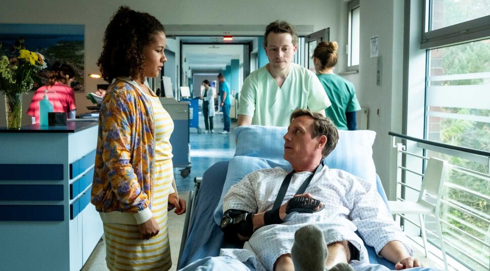 Dr. Moritz Neiss liegt in einem Krankenhausbett in seinem OP-Hemd und wird von Pfleger zum OP geschoben.