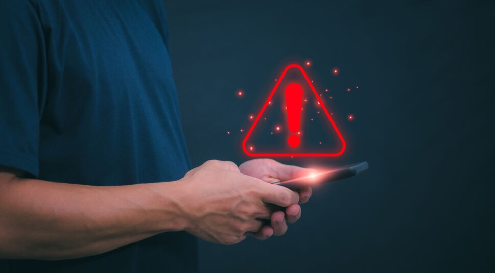 Auf diesem Bild ist eine Person zu sehen, die ihr Smartphone in beiden Händen hält, während über dem Display ein rotes Warnsymbol mit Ausrufezeichen eingeblendet ist – eine Illustration dafür, dass du dein Handy abkühlen solltest, sobald eine Warnmeldung auf eine zu hohe Temperatur hinweist.