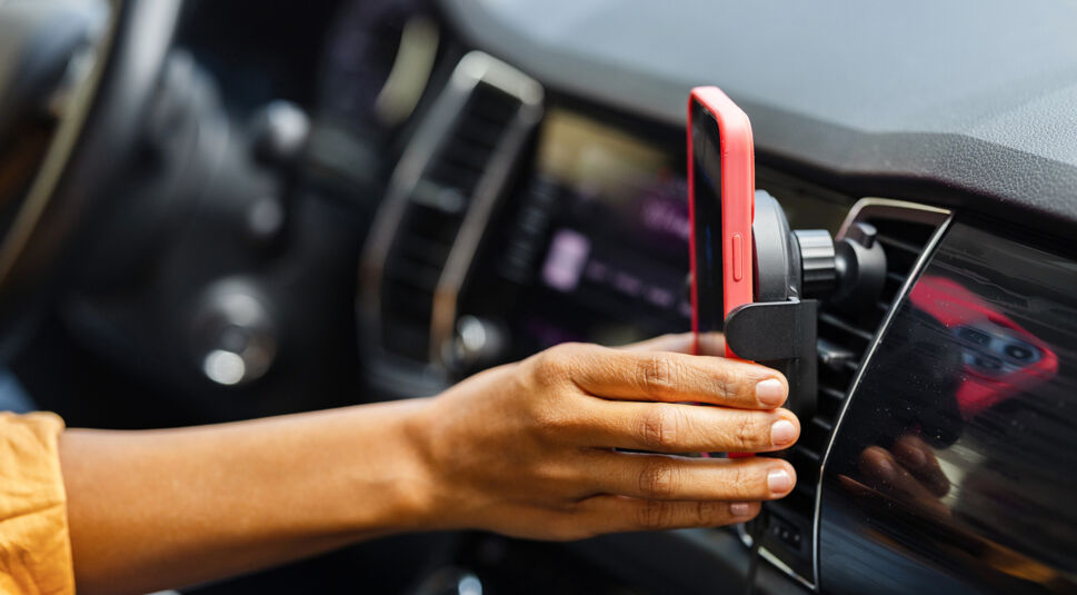 Auf diesem Bild ist eine Hand zu sehen, die ein rotes Smartphone in eine Handyhalterung am Lüftungsschlitz im Auto steckt – ein Ort, an dem das Handy heiß werden kann, wenn direkte Sonne und Hitze im Fahrzeuginnenraum zusammenkommen.