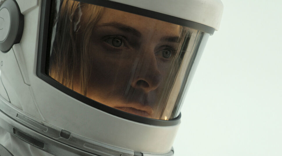 Das Bild zeigt Rebecca Ferguson in einer Nahaufnahme aus Silo Staffel 2. Sie trägt einen weißen, hochtechnologischen Helm, dessen großes Visier einen klaren Blick auf ihr Gesicht ermöglicht. Ihre Augen sind ernst und fokussiert, und man kann eine leichte Besorgnis in ihrem Blick erkennen. Das Bild konzentriert sich auf ihre Augen und den oberen Teil ihres Gesichts, betont durch die Reflektionen im Visier, die eine intensive und dramatische Atmosphäre schaffen. Der Hintergrund ist unscharf, was den Fokus weiter auf ihr Gesicht lenkt und die Isolation oder die Schwere der Situation, in der sie sich befindet, unterstreicht.