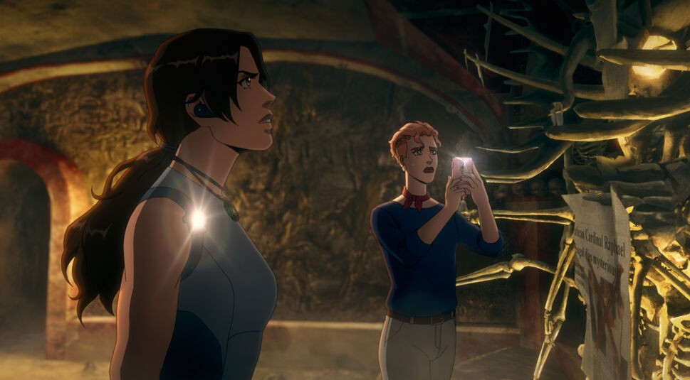 Auf dem Bild aus Tomb Raider: The Legend of Lara Croft sind Lara Croft und Camilla Roth in einer mysteriösen Höhle zu sehen. Lara trägt ein blau-graues Outfit und eine Lampe, die ihren entschlossenen Blick erhellt. Neben ihr steht Camilla Roth, die ein Smartphone in der Hand hält und etwas fotografiert, während sie besorgt wirkt. Die Umgebung ist düster und voller antiker, bedrohlicher Details, was die geheimnisvolle Atmosphäre verstärkt.