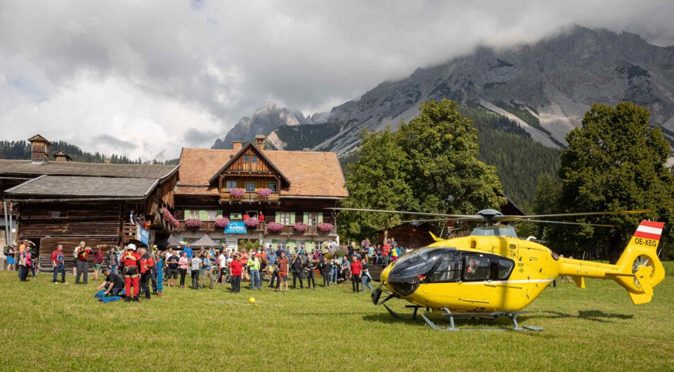 Landschaftsaufnahme mit einem alten Bauernhaus, einem gelben Rettungshubschrauber und Bergen im Hintergrund