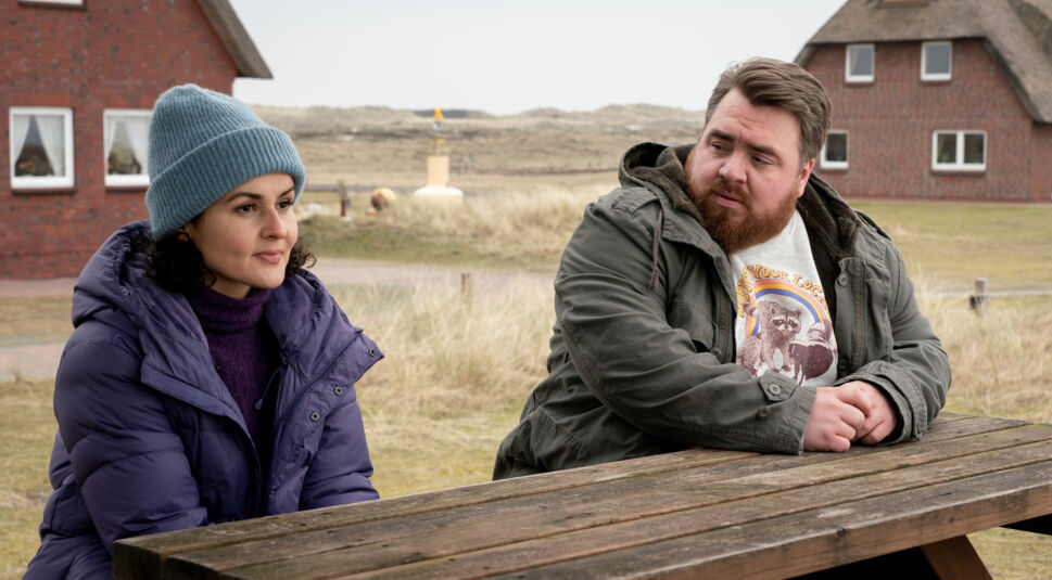 Die Schauspieler Nagmeh Alaei und Tristan Seith sitzen im Freien an einem Holztisch.