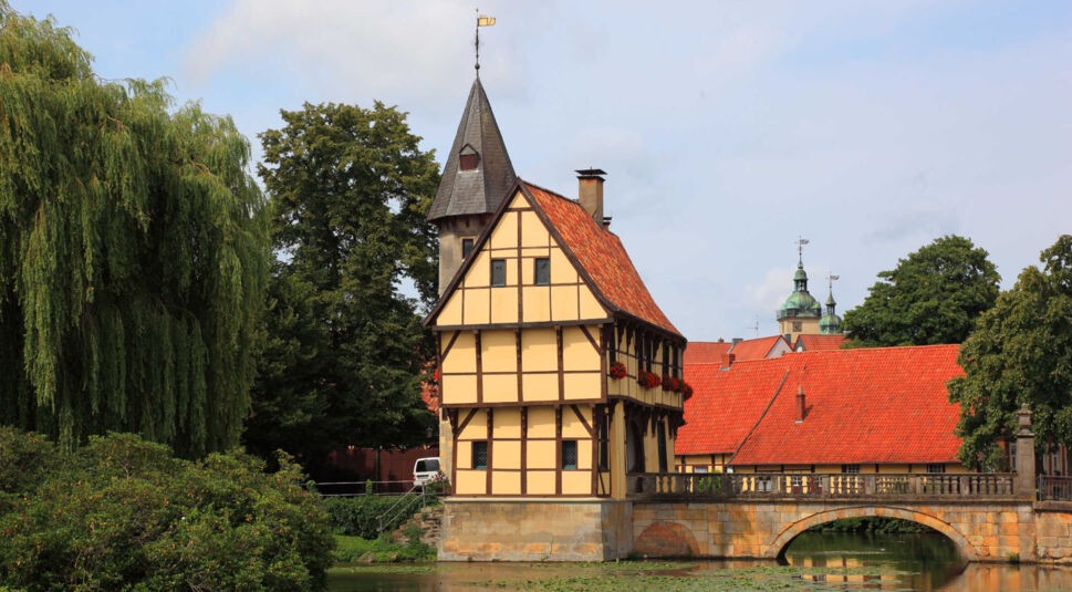 Ein gelbes Fachwerkhaus vor einem See mit Seerosen, dahinter eine dunkle Turmspitze