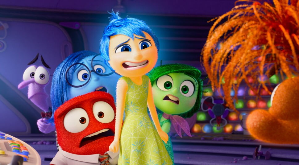 Auf dem Bild aus dem Artikel zu "Alles steht Kopf 3" sind die Charaktere Angst, Kummer, Wut, Freude, Ekel und ein neuer orangefarbener Charakter zu sehen. Angst ist lila, Kummer blau, Wut rot, Freude gelb, und Ekel grün. Alle blicken schockiert oder besorgt in Richtung des orangefarbenen Charakters mit buschigem Haar, der rechts im Bild zu sehen ist. Die Szene spielt in einer farbenfrohen, futuristischen Umgebung.