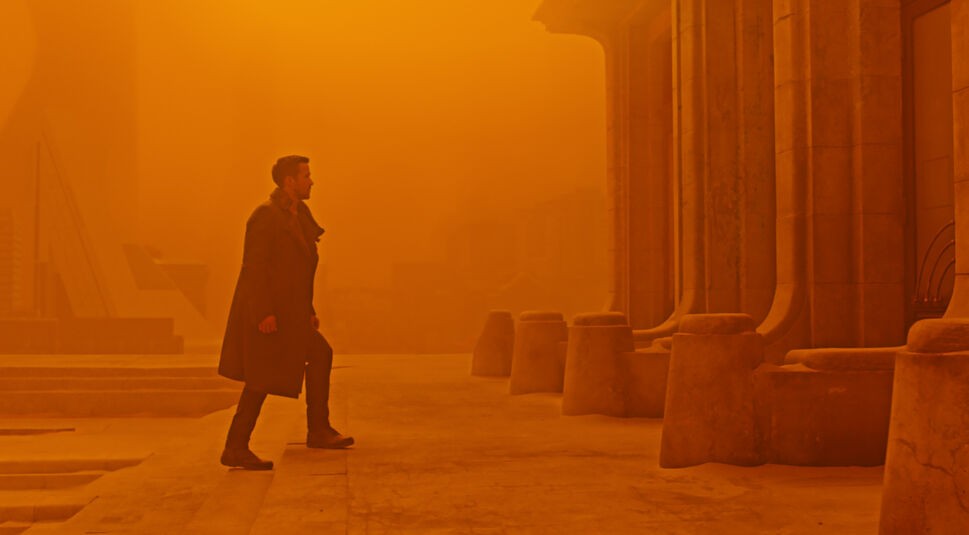 Dieses Bild zum Blade Runner 2099-Artikel stammt aus Blade Runner 2049 und zeigt Ryan Gosling, wie er eine Treppenstufe aufsteigt während das Bild orange eingefärbt ist.