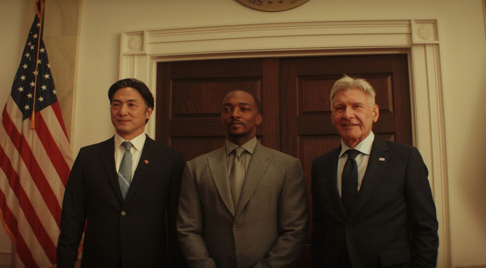 Auf dem Bild im Artikel zu Captain America 4: Brave New World sind drei Personen von vorne zu erkennen. Sie stehen nebeneinander, lächeln und schauen geradeaus an der Kamera vorbei. Hinter ihnen ist eine weiße Wand mit einer braunen massiven Holztür. Am linken Bildrand ist die Flagge der Vereinigten Staaten zu sehen, die neben der Türaufgehängt wurde. Die Person links ist asiatisch und trägt einen schwarzen Anzug mit weißem Hemd und blauer Krawatte. In der Mitte steht Captain America. Er ist schwarz. Er trägt einen grauen Anzug mit grauem Hemd und grauer Krawatte. Neben ihm, rechts im Bild steht ein weißer Mann mit grauen Haaren, einem schwarzen Anzug, weißem Hemd und schwarzer Krawatte. Es ist Harrison Ford, der im Film den Präsidenten der USA spielt.