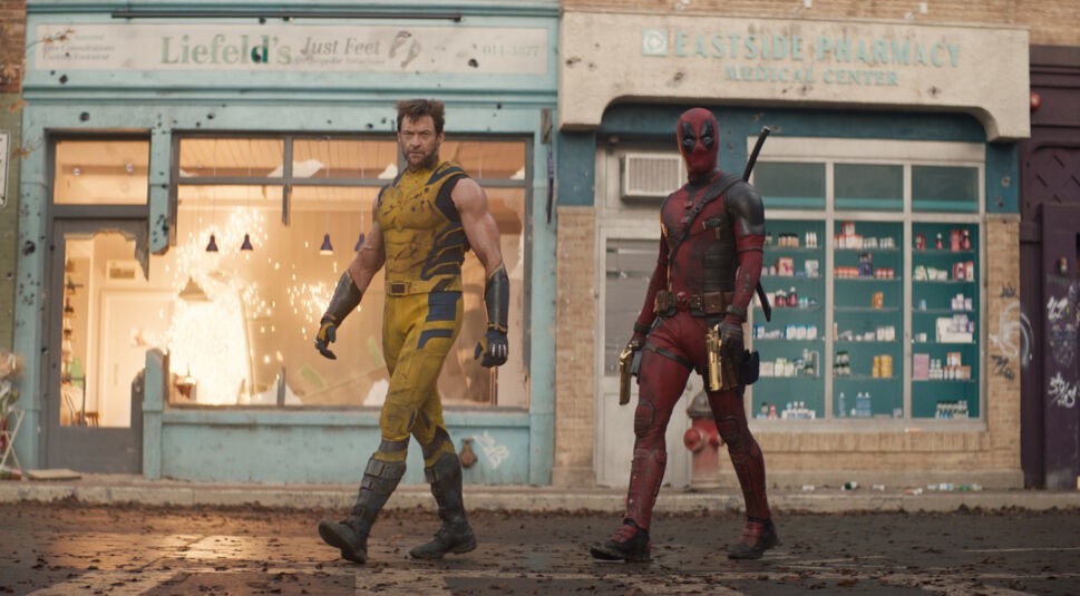 Auf dem Bild zu "Deadpool & Wolverine Ende erklärt" sind die Charaktere Deadpool und Wolverine in einer dramatischen Szene zu sehen. Links steht Wolverine, gespielt von Hugh Jackman, in einem gelben Kampfanzug mit ernstem Blick und muskulösem Auftreten. Rechts von ihm steht Deadpool, dargestellt von Ryan Reynolds, in seinem ikonischen roten Anzug mit schwarzem Maskengesicht, der eine entspannte Haltung einnimmt. Beide Charaktere gehen eine Stadtstraße entlang, im Hintergrund sind beschädigte Gebäude und eine Apotheke zu sehen. Die Szene vermittelt eine angespannte, actionreiche Stimmung.