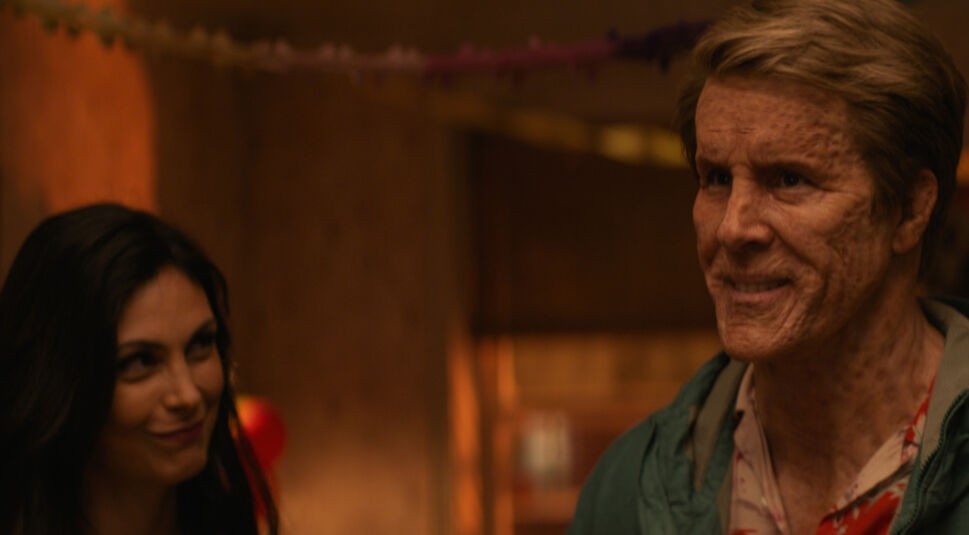 Auf dem Bild zu "Deadpool & Wolverine Ende erklärt" sind Vanessa und Wade Wilson in einer emotionalen Szene zu sehen. Links steht Vanessa, gespielt von Morena Baccarin, mit einem sanften Lächeln auf den Lippen. Rechts von ihr steht Wade Wilson, dargestellt von Ryan Reynolds, mit entstelltem Gesicht und einem leichten, erleichterten Lächeln. Im Hintergrund sind unscharfe Partykulissen zu erkennen, was eine intime und warme Atmosphäre vermittelt.