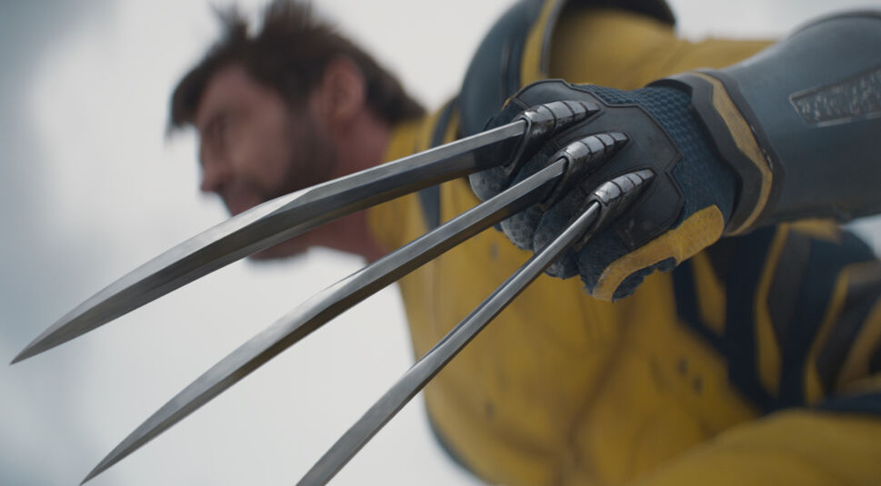Das Bild zeigt eine Nahaufnahme von Wolverine, gespielt von Hugh Jackman, mit Fokus auf seine berühmten Adamantium-Klauen, die auch in Deadpool & Wolverine eine Rolle spielen. Die scharfen, metallischen Klauen sind ausgefahren und dominieren den Vordergrund des Bildes, während Wolverines Gesicht und Oberkörper im Hintergrund leicht unscharf zu sehen sind. Er trägt seinen gelb-blauen Anzug, der an das klassische Comic-Kostüm erinnert. Die Szene vermittelt eine intensive und bedrohliche Stimmung, passend zu Wolverines kämpferischem Charakter und seinen beeindruckenden Waffen.