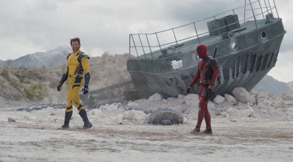Das Bild aus der Deadpool & Wolverine-Kritik zeigt eine Szene mit Deadpool (Ryan Reynolds) und Wolverine (Hugh Jackman), die zusammen durch eine karge, felsige Landschaft gehen. Im Hintergrund befindet sich ein großes, verrostetes Schiffswrack, das auf einem trockenen, wüstenartigen Boden liegt. Die Umgebung wirkt trostlos und öde, mit wenigen Pflanzen und kahlen Hügeln im Hintergrund.