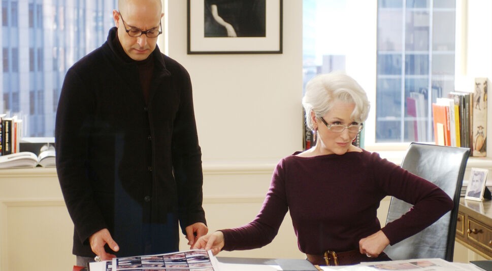 In Der Teufel trägt Prada sitzt die Chefredakteurin Miranda Priestly, gespielt von Meryl Streep, an ihrem Schreibtisch. Neben ihr steht in der linken BIldhälfte ihr Art Director. Auf dem Schreibtisch vor ihnen liegt ein Ausdruck von einigen Bildern, über den sie gerade sprechen. Miranda stützt ihren Arm an ihrer Hüfte ab und schaut über ihre Brille hinweg nach vorne, vermutlich zur Tür ihres Büros. Im Hintergrund sieht man zwei Fenster und die Fassade von angrenzenden Hochhäusern in New York.