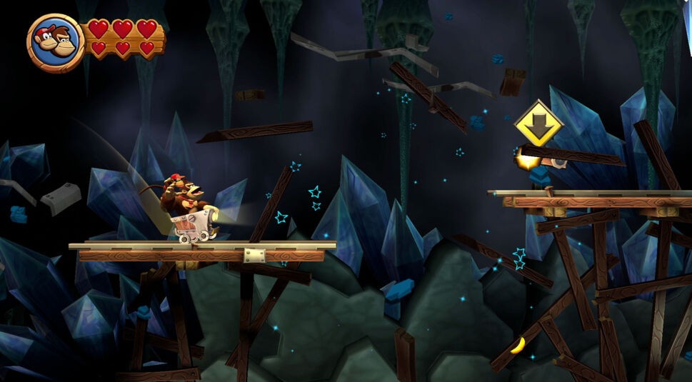 Auf dem Bild aus Donkey Kong Country Return HD ist Donkey Kong in einer Lore zu sehen, die auf Schienen fährt. Die Umgebung ist eine dunkle Höhle mit blauen Kristallen und Holzplanken. Über Donkey Kong und Diddy Kong ist eine Herzleiste mit vier Herzen. Sie scheinen bereit zu sein, über eine Lücke in den Schienen zu springen. Ein gelber Pfeil nach unten deutet auf eine kommende Aktion hin.