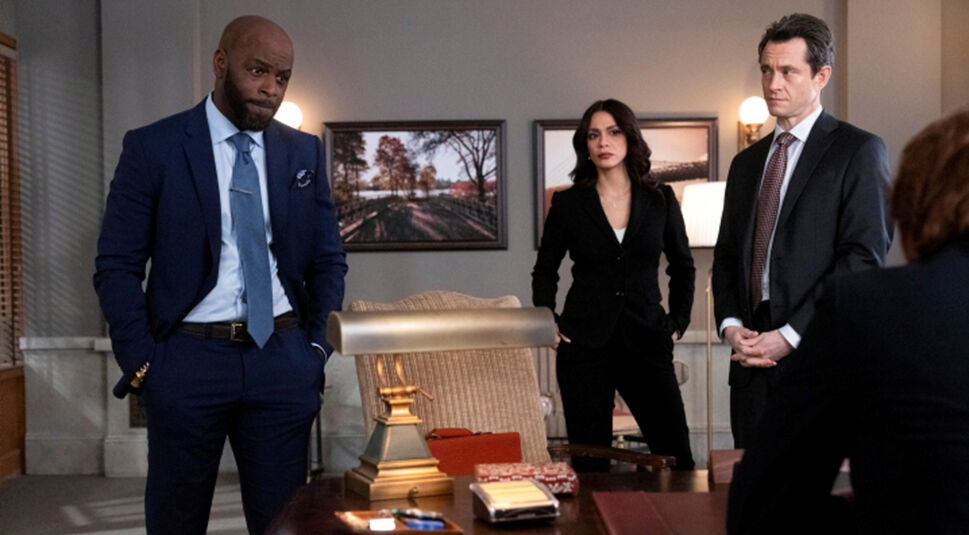 Das Bild im Artikel zu den GigaTV-Highlights im Juli 2024 zeigt drei Personen in einem Büro in der Staffel 4 von Law & Order: Organized Crime, die ernst aussehen. Links steht ein Mann mit Glatze in einem blauen Anzug und Krawatte, der die Hände in die Hosentaschen gesteckt hat. In der Mitte steht eine Frau mit langen, dunklen Haaren in einem schwarzen Anzug, die ihre Hände ebenfalls in den Hosentaschen hat. Rechts steht ein Mann mit dunklem Haar in einem dunklen Anzug und gestreifter Krawatte, der seine Hände vor sich gefaltet hat. Sie stehen um einen Schreibtisch, auf dem eine Lampe, Bücher und einige Büromaterialien liegen. Im Hintergrund hängen gerahmte Bilder an der Wand.