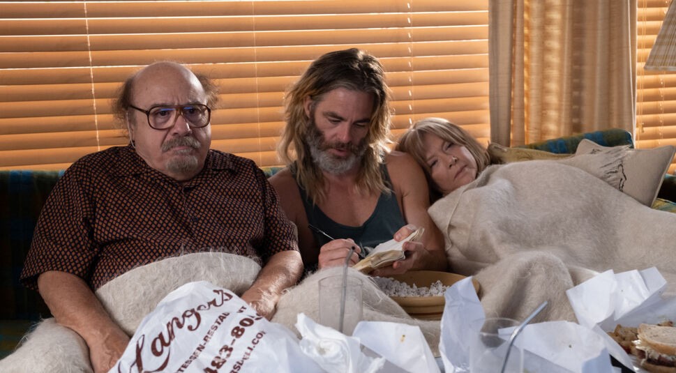 Das Bild im Artikel zu den GigaTV-Highlights im Juli 2024 zeigt drei Personen, die auf einem Sofa sitzen und in Decken gehüllt sind. Links sitzt Danny DeVito als Jack. In der Mitte sitzt Chris Pine als Darren mit langen blonden Haaren und Bart, der in einem Notizbuch schreibt. Rechts lehnt Jennifer Jason Leigh als Susan an seiner Schulter und scheint zu schlafen. Vor ihnen auf dem Tisch liegen Essensverpackungen und eine Schüssel mit Popcorn. Im Hintergrund sind geschlossene Jalousien vor einem Fenster zu sehen.