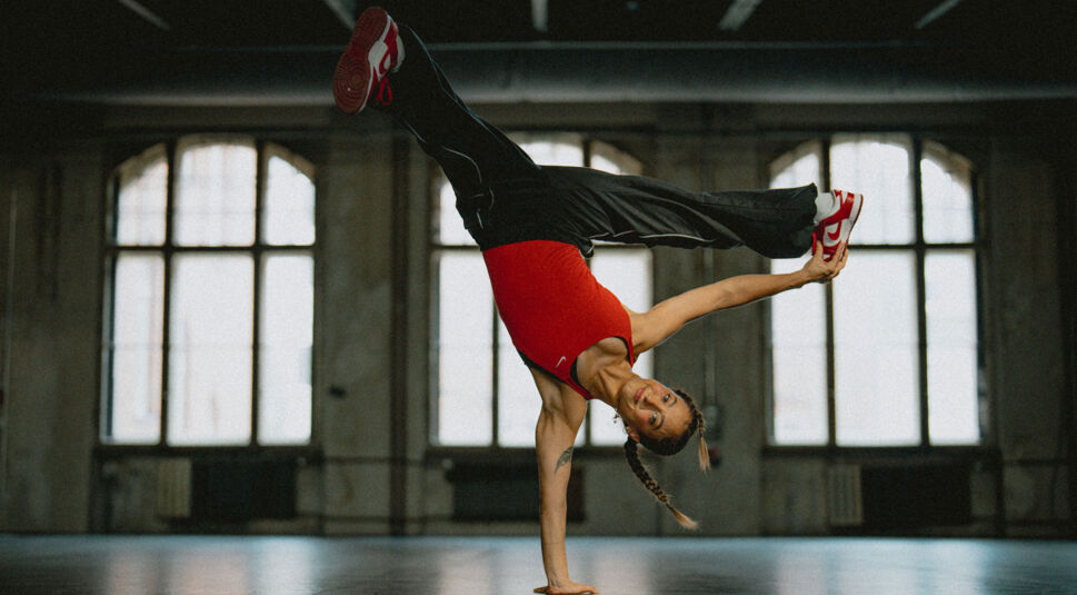 Auf diesem Bild ist die Breakerin Jilou Rasul für die neue Vodafone GO Werbung zu sehen. Sie macht einen Breakdance-Move, indem sie sich auf einer Hand abstützt und ihr Körper in der Luft ist. Mit der linken Hand berührt sie ihren Fuß. Sie trägt eine schwarze Jeans und ein rotes Top.
