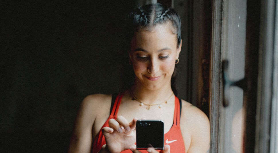 Jilou Rasul schaut auf ein Samsung Galaxy Z Flip mit einem zufriedenen Gesichtsausdruck. In der GO Werbung 2024 von Vodafone zeigt sie, wie sie zum Breaken gekommen ist und was ihr GO-Moment war. Passend dazu trägt sie ein rotes Sportoberteil.