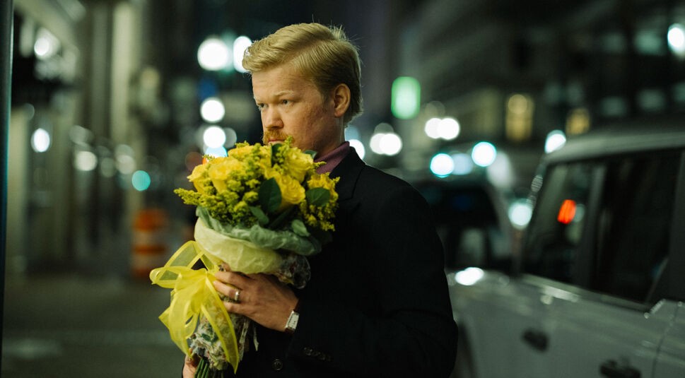 Auf dem Bild des Kinds of Kindness-Ende-erklärt-Artikels trägt Jesse Plemons einen dunklen Anzug und ein violettes Hemd. Er hält einen großen, gelb umwickelten Blumenstrauß, während er nachdenklich zur Seite schaut. Die Szene spielt nachts, erkennbar an den unscharfen Lichtern und der städtischen Umgebung im Hintergrund.