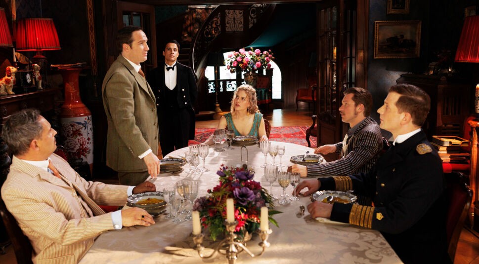 In dieser Szene aus der Prime-Video-Serie Miss Sophie sitzt Miss Sophie (Alicia von Rittberg) mit ihren illustren Gästen – Mr. Pommeroy (Moritz Bleibtreu), Mr. Winterbottom (Frederick Lau), Sir Toby (Jacob Matschenz) und Admiral von Schneider (Christoph Schechinger) – an einer reich gedeckten Tafel. Butler James (Kostja Ullmann) steht im Hintergrund. Die Stimmung ist festlich und nostalgisch.