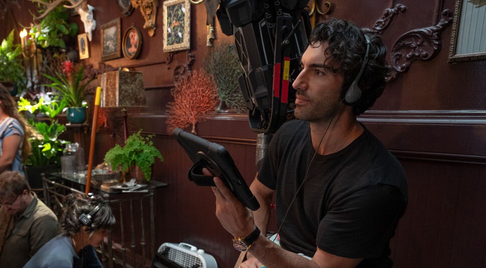 Schauspieler und Regisseur Justin Baldoni ist am Set des Films Nur noch ein einziges Mal. Er sitzt in einem dunken, rustikal eingerichteten Raum, vermutlich ein Restaurant. Er trägt Kopfhörer und in seiner Hand hält er ein Tablett. Er schaut konzentriert vor sich. Auf dem Bild ist nicht zu sehen, wen oder was er ansieht, aber vermutlich wird gerade eine Szene des Films gedreht und er betreut den Vorgang als Regissseur. Am linken Bildrand sind zwei weitere Personen zu sehen, die vermutlich zur Film-Crew gehören.