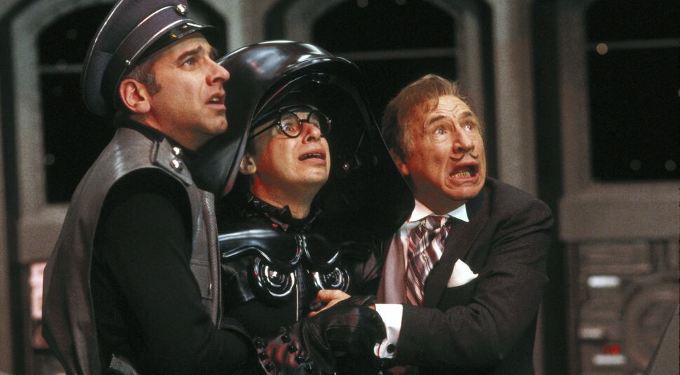 Ein Szenenbild aus Spaceballs zeigt Lord Helmchen mit zwei Männern links und rechts von ihm. Die drei scheinen sich aneinander festzuhalten und schauen entsetzt nach rechts oben, als würde von dort etwas auf sie zufliegen. Der Mann rechts hat eine Art Uniform an, wie ein Schaffner oder Polizist. Lord Helmchen hat eine schwarz glänzende Uniform an, die an Darth Vader erinnern soll. Sein Helm ist viel zu groß und darunter trägt er eine runde, dickrandige Brille. Der Mann links trägt Anzug und Krawatte. In sein Gesicht ist ein Schnurrbart gemalt..