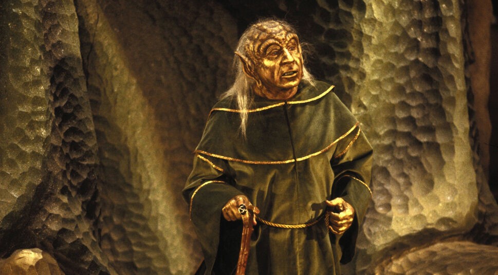 Ein Szenenbild aus Spaceballs zeigt Mel Brooks in der Rolle von Joghurt. Er steht zentral im Bild mit gold-grünem Umhang vor einer Höhlenwand, die in ähnlichen Farben schimmert. Sein Gesicht ist golden und faltig. Sein Schädel ist größer als der eines Menschen und seine Ohren spitz. Über die Schultern fällt graues, dünnes Haar.