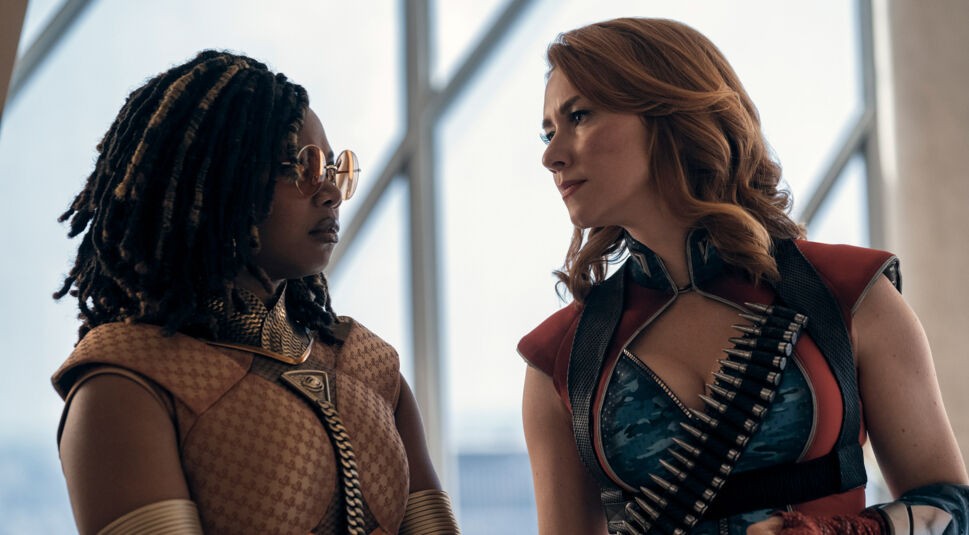 Auf diesem Bild zum The Boys Staffel 5 Ausblick siehst Du zwei Frauen, die sich intensiv und vielleicht sogar herausfordernd anschauen. Links steht Sister Sage, gespielt von Susan Heyward, in einer auffälligen, stilvollen Kampfausrüstung mit goldfarbenen Details und Sonnenbrille. Rechts ist Firecracker zu sehen, dargestellt von Valorie Curry, in einem rot-blauen Kostüm mit einer Brille und Patronengurten. Beide Charaktere wirken stark und entschlossen, und ihre Kleidung deutet auf eine actionreiche Szene hin.