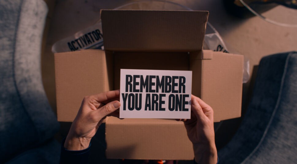Auf dem Bild aus dem The Substance-Film ist ein brauner Karton von oben zu sehen. Er steht offen. Darüber halten zwei Hände eine Postkarte auf der in großen schwarzen Buchstaben auf weißem Hintergrund steht: "REMEMBER YOU ARE ONE"
