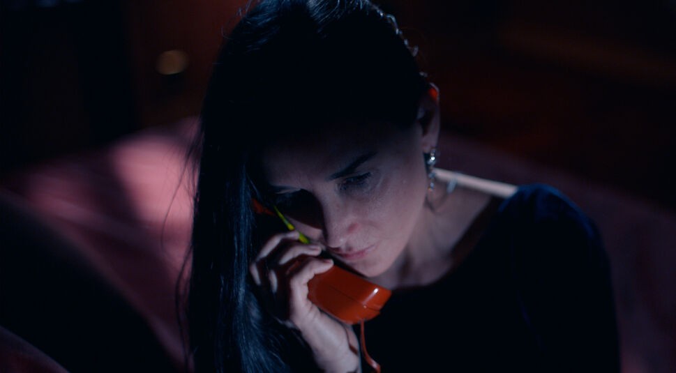 Das Bild aus dem Film The Substance zeigt Demi Moore in einer Nahaufnahme. Sie hat das Gesicht nach unten gewandt und hält einen roten Telefonhörer an ihr Ohr. Ihre dunklen Haare fallen über ihre Schulter und das Telefon nach unten. Um sie herum ist es dunkel im Raum.